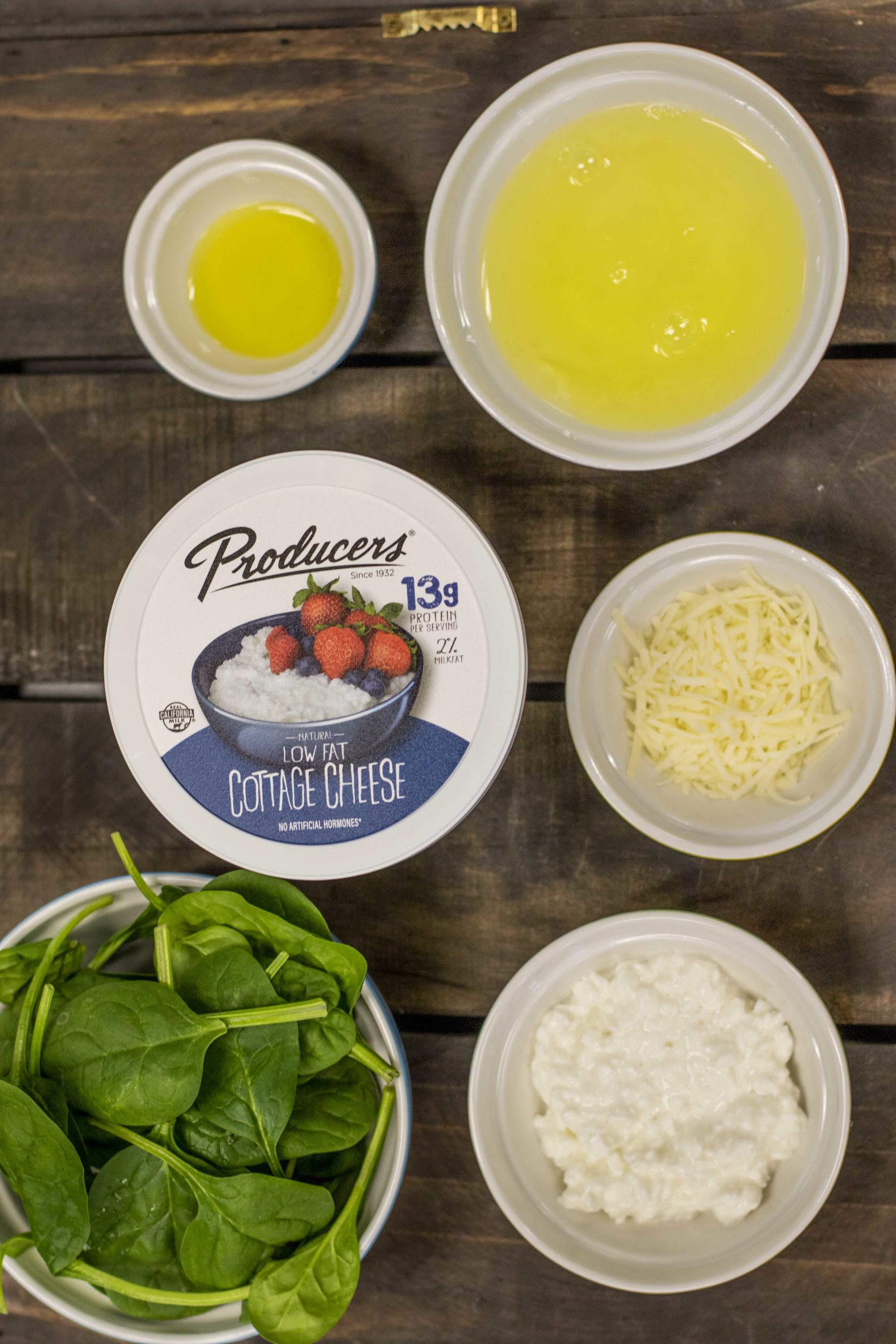 ingredients Producers Low Fat Cottage Cheese surrounded by spinach, oil, egg whites, and cheese on a wood table.