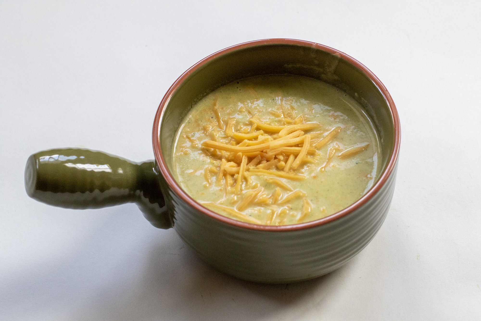 Broccoli Soup in a green bowl topped with shredded cheddar cheese laying on a flat white background.
